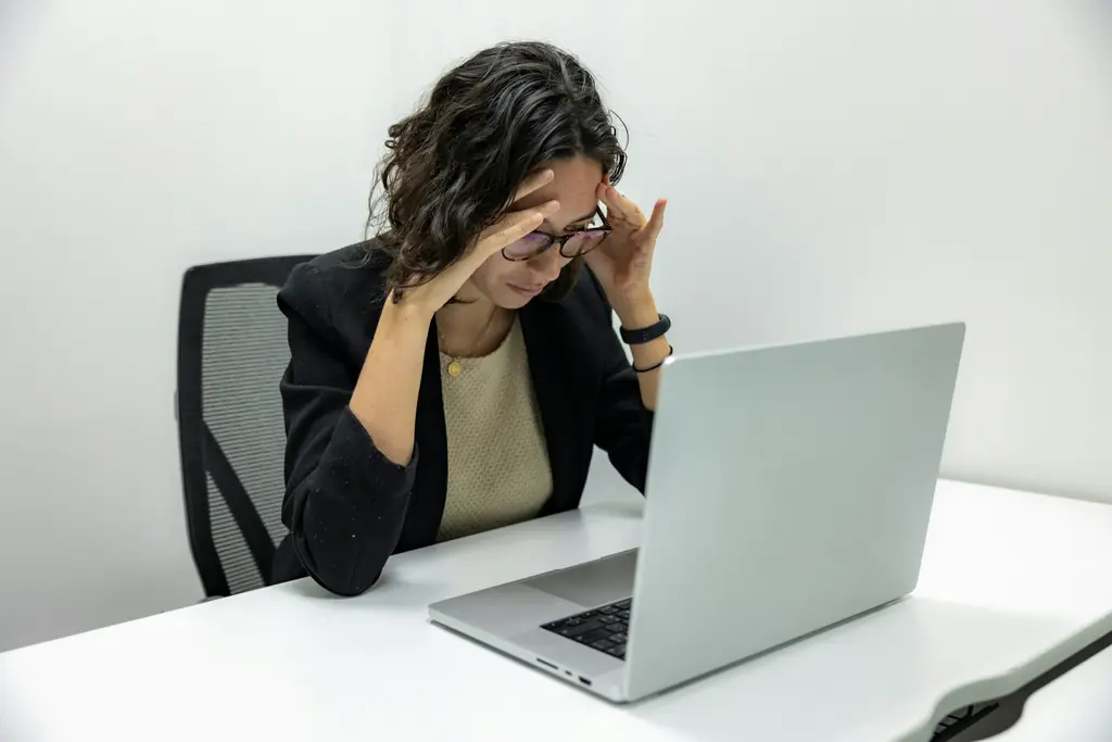 A woman at a desk holding her head beside a laptop