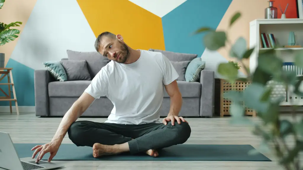 A man doing a seated stretch on a mat in front of a laptop at home