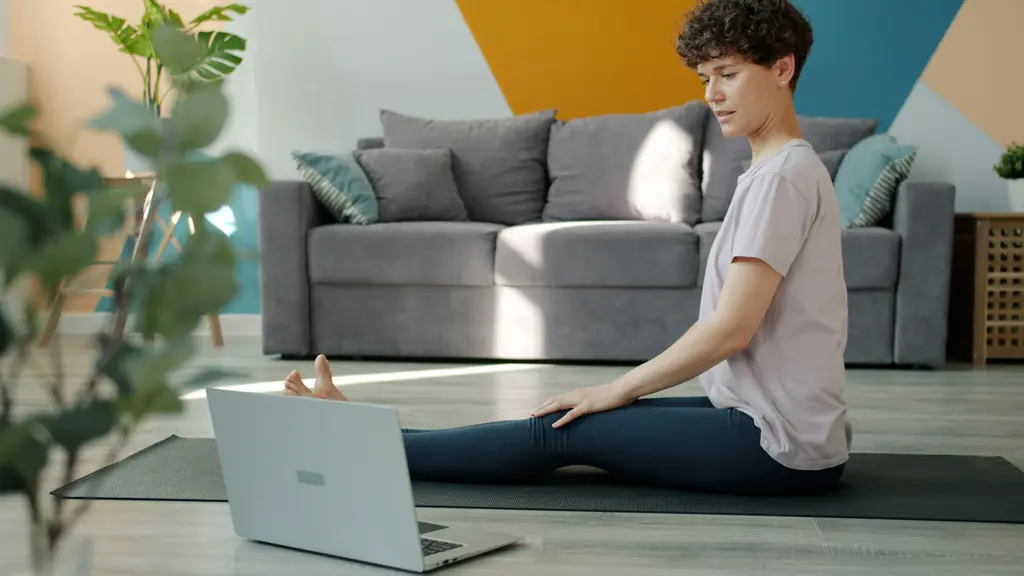 A woman sitting on an exercise mat in front of a laptop at home