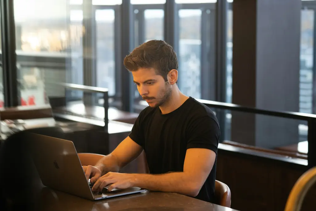 A man typing on a laptop at a table by a window