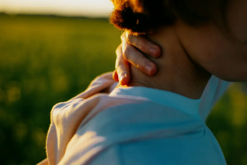 A person holding the side of their neck and shoulder outdoors