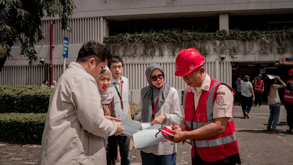 A worker in a red hard hat and vest talking with a small group outdoors